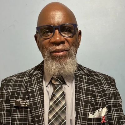 Elderly man in glasses and plaid suit with name tag and beard.