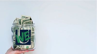 Hand holding a jar full of dollar bills with 'WE' written on it.