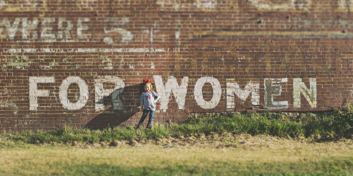 A young girl stands confidently against a brick wall with faded graffiti that reads 'FOR WOMEN'.