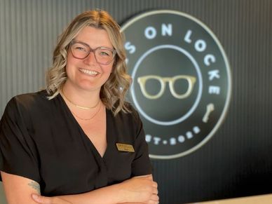 Smiling woman with glasses in front of an eyewear store sign.