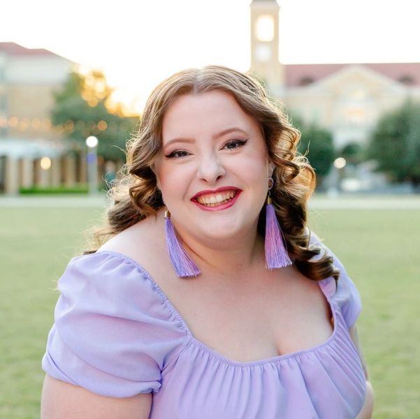 Smiling woman in a light purple dress outdoors with matching tassel earrings.
