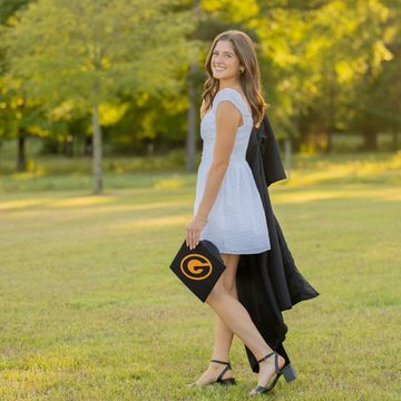 Young woman in white dress holding a graduation cap outdoors.