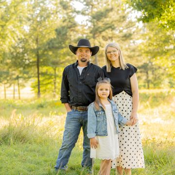 A family of three posing outdoors in a sunny, green field.