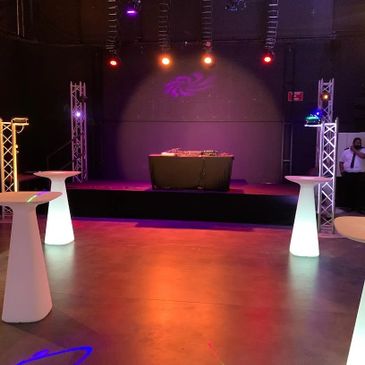 Empty event space with lit cocktail tables and a DJ booth on stage.