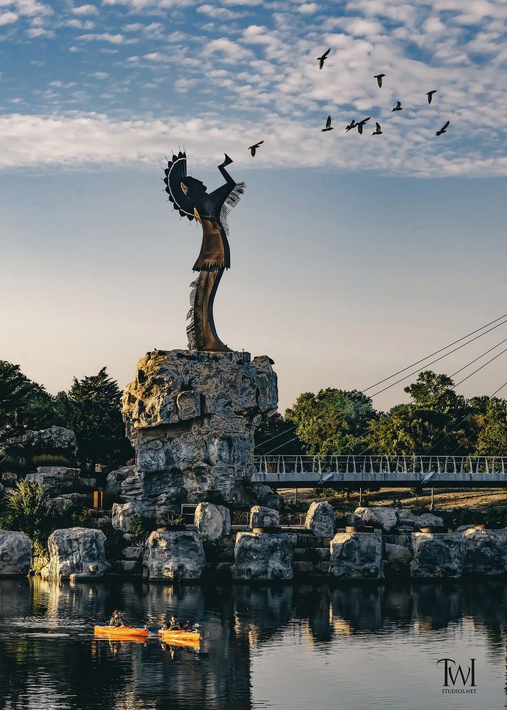 Statue of a Native American figure with birds flying nearby and kayakers on the water.