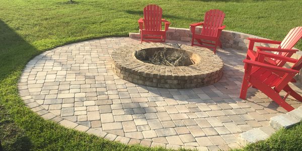 A circular fire pit with pavers around it and 4 red Adirondack chairs posed ready to seat people.