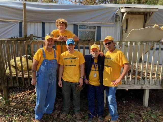 Five men in yellow shirts posing outside a wooden deck and trailer.