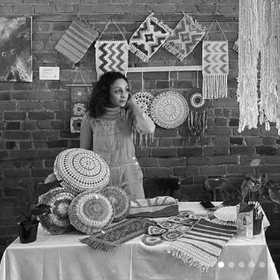 A woman stands behind a table with woven crafts and wall hangings.