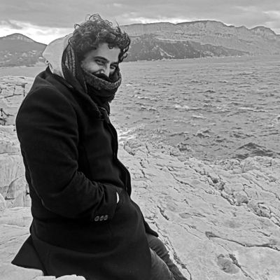 Man bundled up sitting on rocky shore with mountains in background.