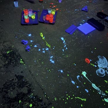 Paint splatters and cups under blacklight on a dark floor.