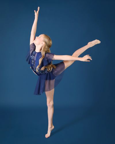 A young dancer in a blue dress performs an elegant arabesque against a blue backdrop.