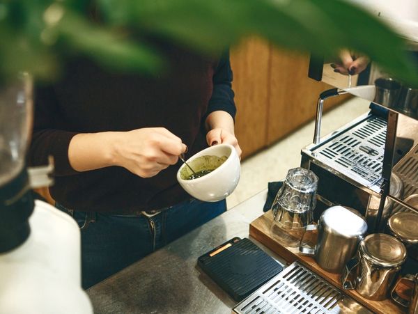 Barista stirring a cup of green tea or matcha beside coffee equipment.