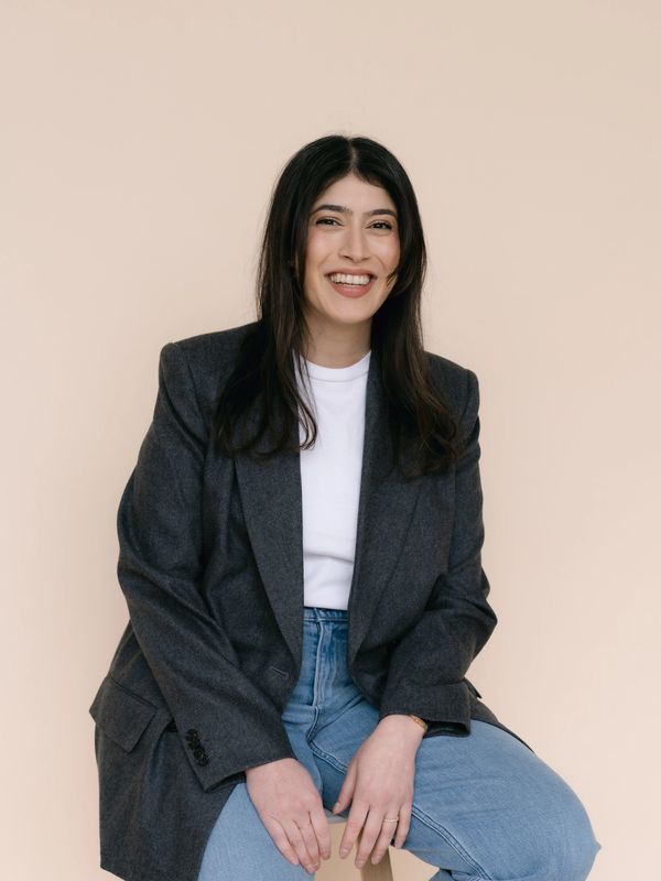 Young woman smiling, seated on a stool against a beige background.
