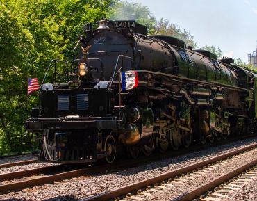 A vintage steam locomotive with American flags on the front, traveling on railway tracks surrounded by greenery.