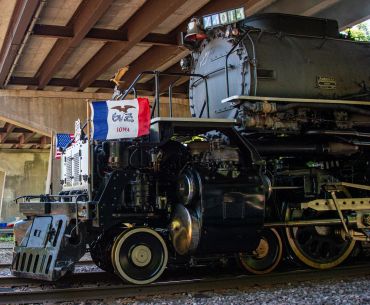 A vintage steam locomotive with an Iowa flag under a bridge.