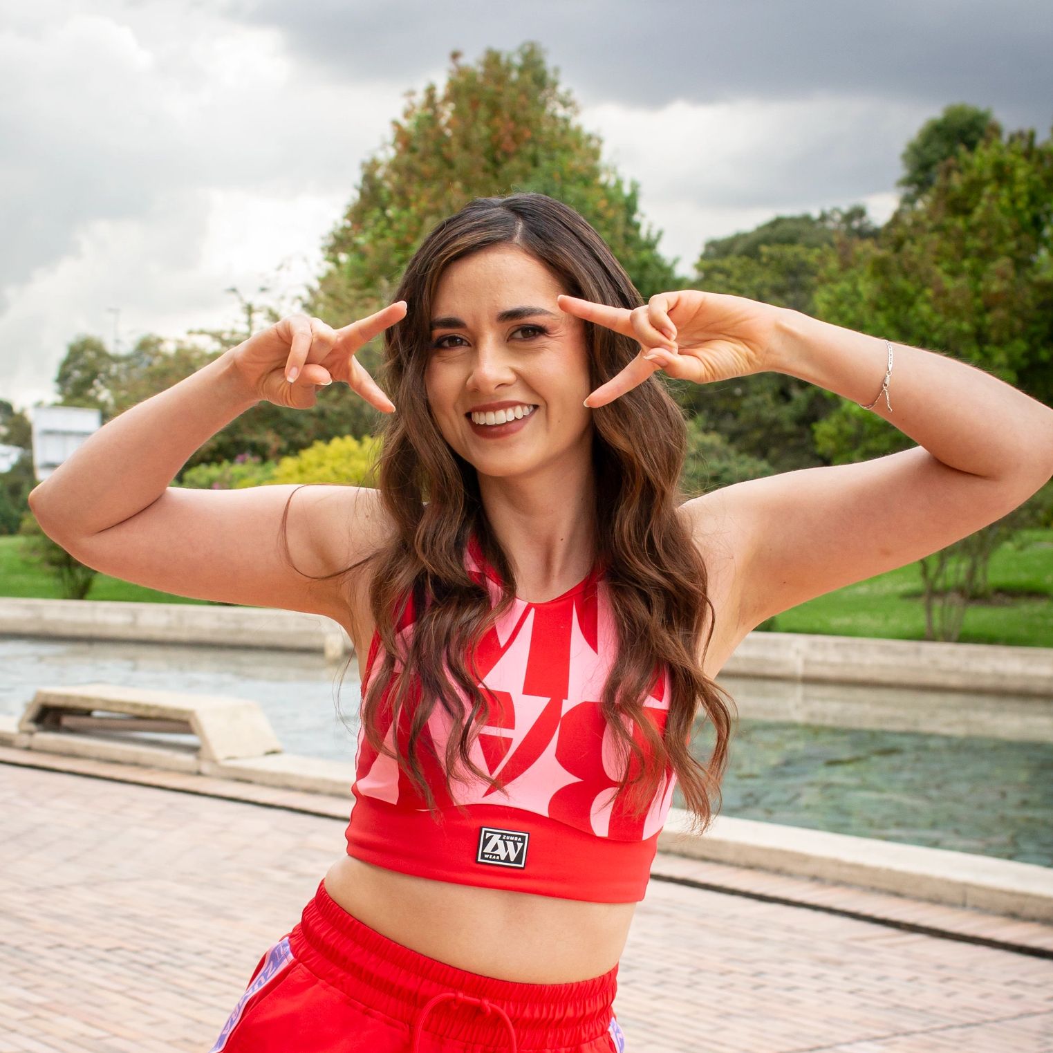 Smiling woman in red workout clothes making a playful peace sign gesture outdoors.