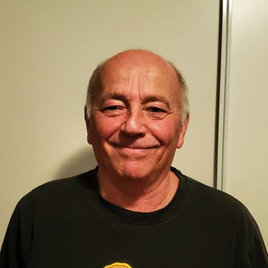 Smiling elderly man in a black shirt against a plain background.