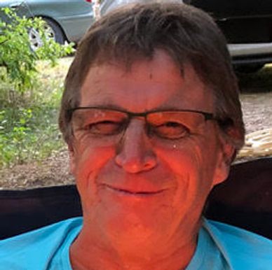 Smiling man wearing glasses outdoors with cars and greenery in background.