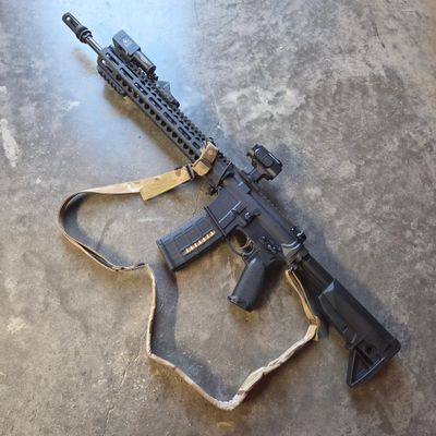 Black tactical rifle with a tan sling on concrete floor.