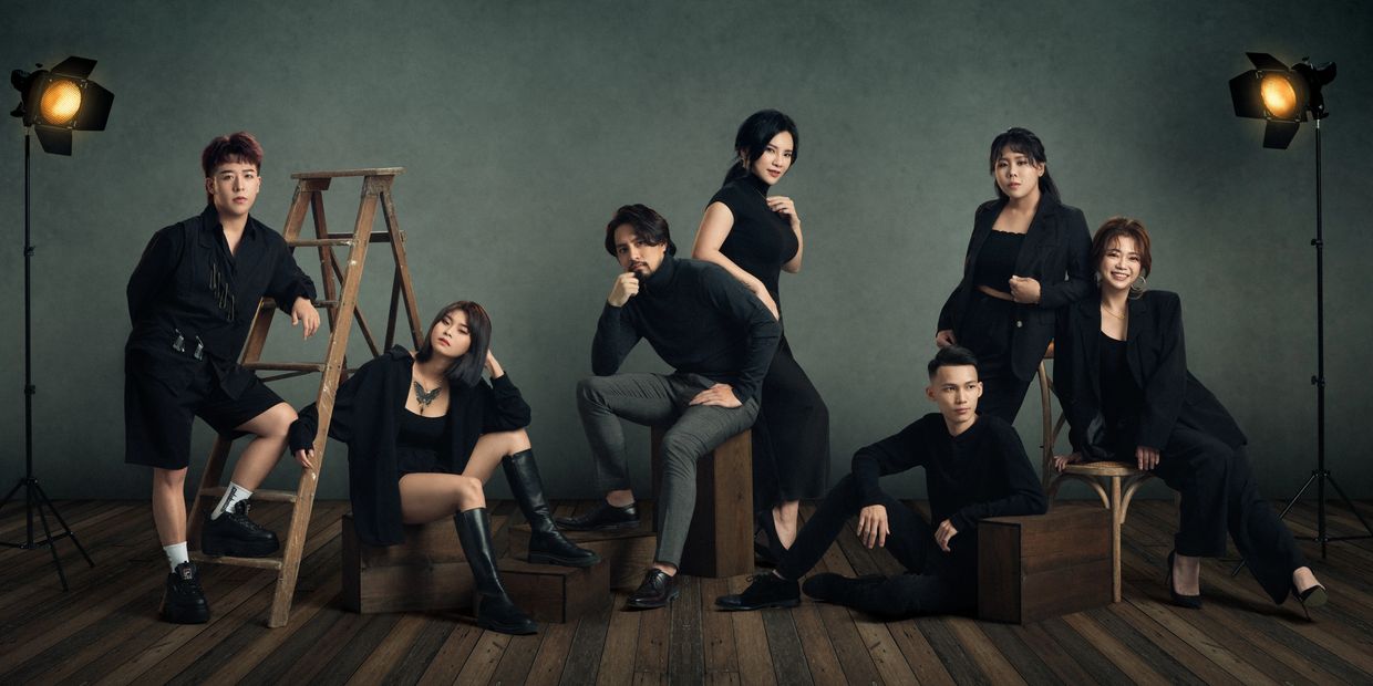 Seven stylish people posing in a moody studio with vintage lighting and wooden floor.