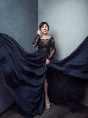 Elegant woman in a flowing black gown posing gracefully against a corner wall.