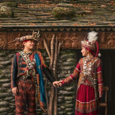 Couple in traditional attire holding hands in front of a decorated stone wall.
