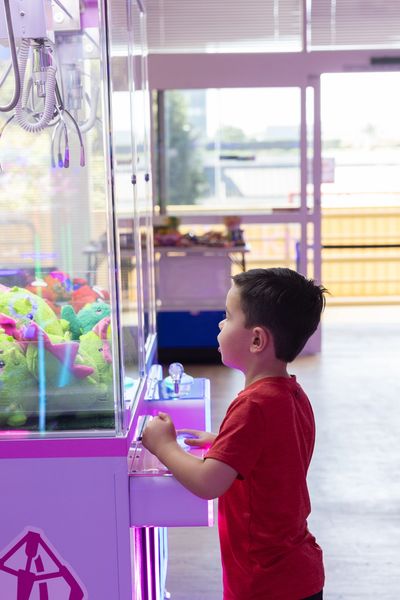 A young boy in a red shirt playing a claw machine filled with colorful stuffed toys.