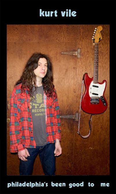 Kurt Vile standing with a red electric guitar hanging on a wooden wall.