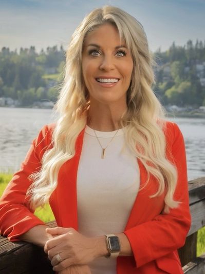 Smiling woman in red blazer by a lakeside with long blonde hair.