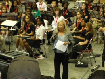 Woman speaking to a seated band or orchestra group.