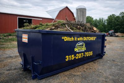 Large dumpster filled with yard debris near a barn.