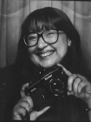 Smiling person holding a Vivitar camera, wearing glasses.