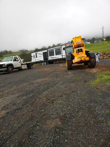 A white truck and yellow JCB loader moving a large trailer on a gravel road.