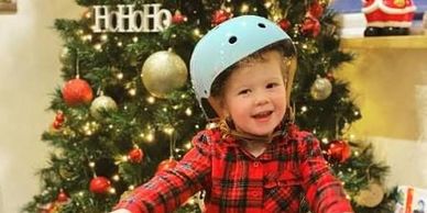 Smiling child wearing a helmet by a decorated Christmas tree.