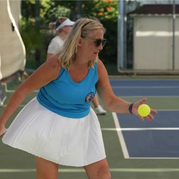 Woman in blue tank top and white skirt playing pickleball with a yellow ball.