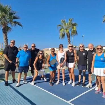 Group of people posing on a pickleball court with paddles, palm trees in the background.