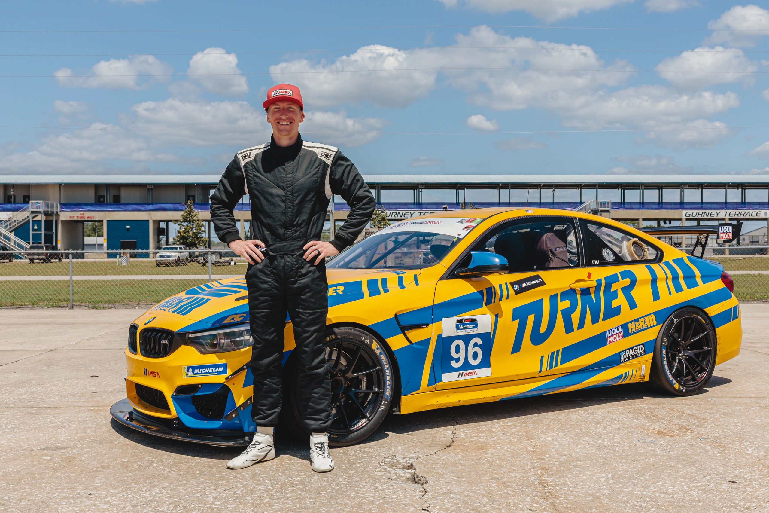 Race car driver in black suit stands next to a yellow and blue Turner Motorsport car.