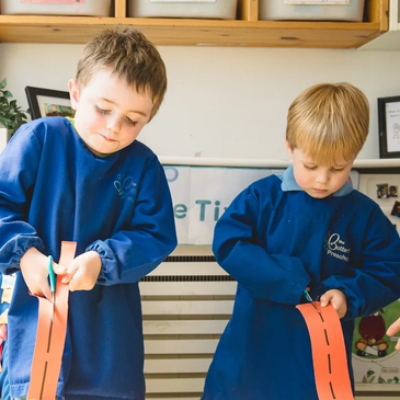 Two young boys in blue aprons cutting orange paper strips with scissors.
