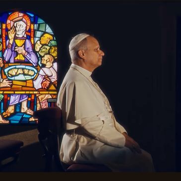 A man in white religious attire sitting beside a colorful stained glass window.