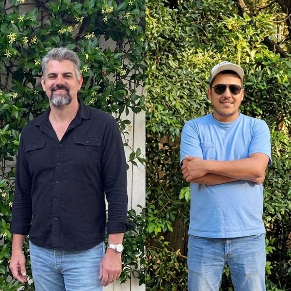 Two men standing outdoors with greenery behind them.