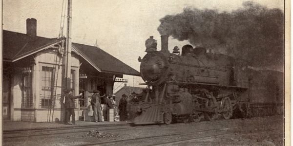 A vintage steam locomotive at a small train station with people waiting in Dixon, MO.