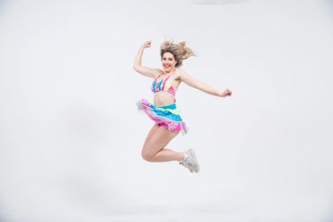 A young woman in colorful dancewear jumps joyfully against a white background.