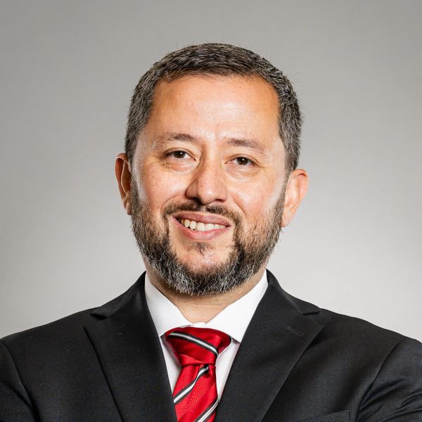 Man in black suit and red striped tie smiling against gray background.