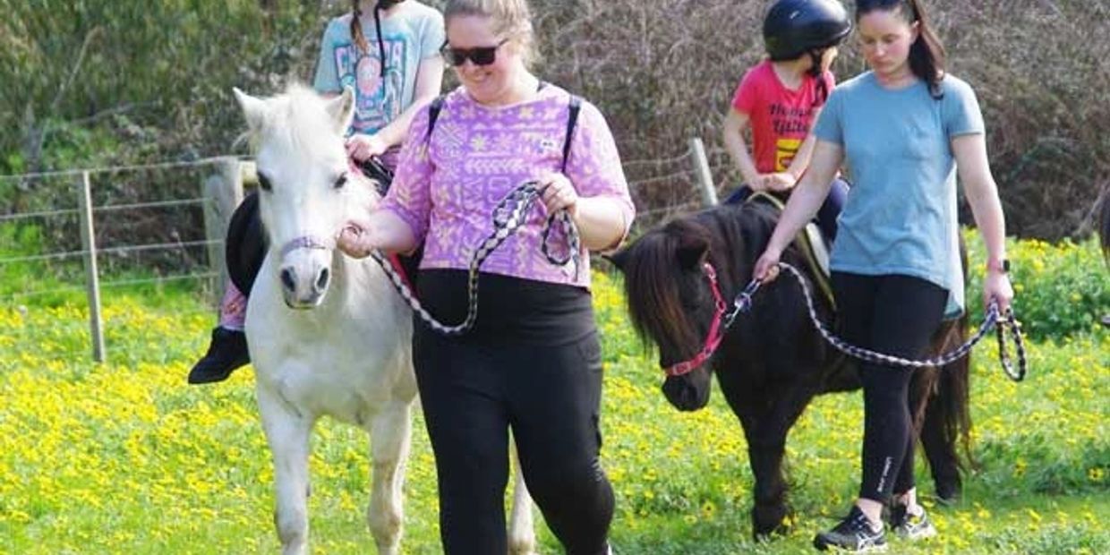Two women leading ponies with kids riding them in a sunny field of yellow flowers.