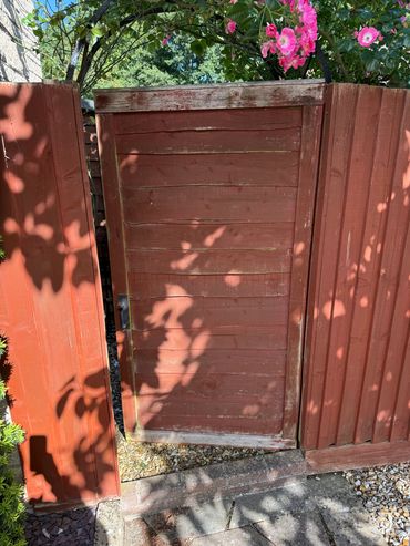 A weathered red wooden gate surrounded by blooming pink flowers and shadows.