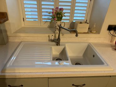 White kitchen sink with dual basins and a black faucet under a window with shutters.