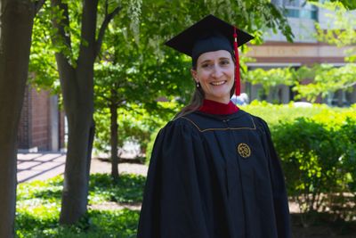 A smiling graduate in cap and gown outdoors on a sunny day.
