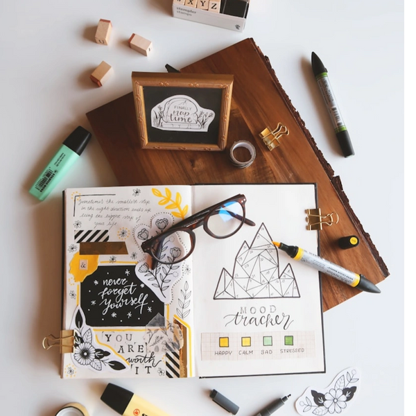 An open mood tracker book with items across the table; highlighter, glasses, photo frame and clips. 