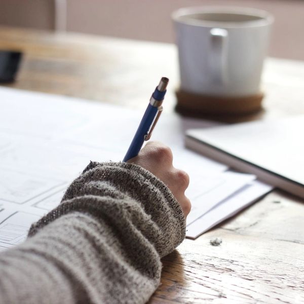 A desk, paper and a hand holding a dark pen, writing on the paper. There is a cup in the background.