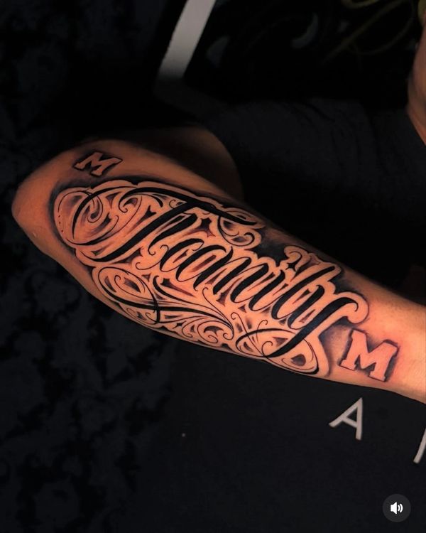 Intricate black ink tattoo on forearm with the word 'Tacaniloj' and decorative flourishes.